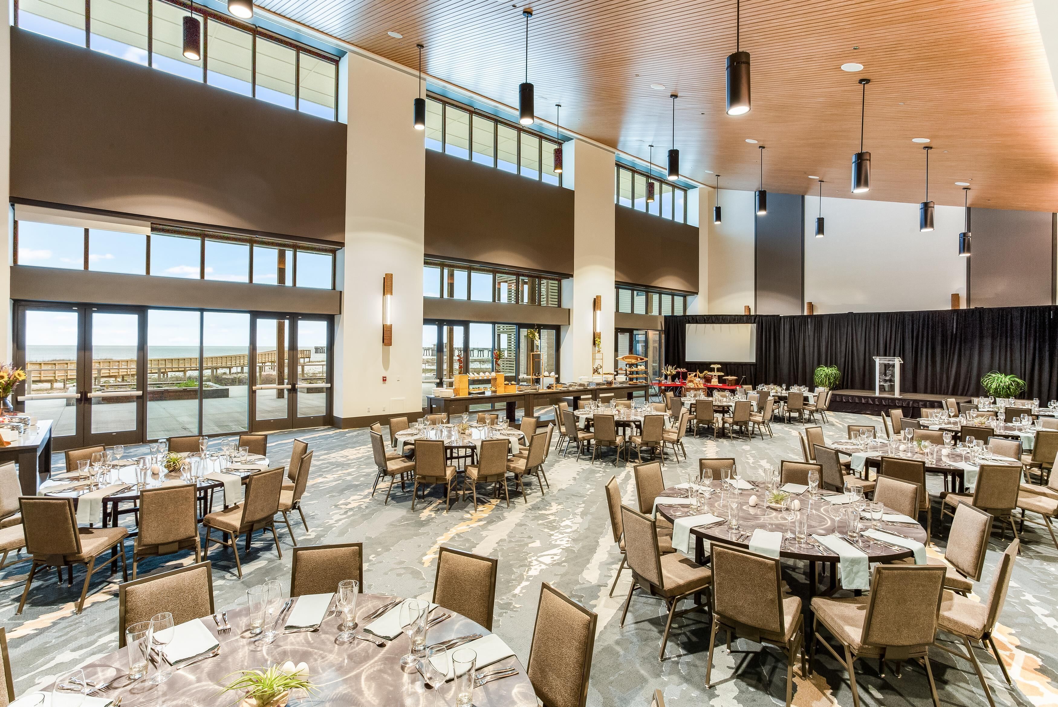 A spacious banquet hall with multiple tables set for dining, large windows offering views, and a stage for presentations.