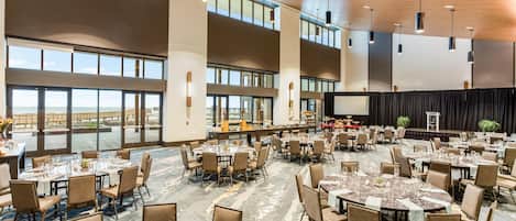 A spacious banquet hall with multiple tables set for dining, large windows offering views, and a stage for presentations.