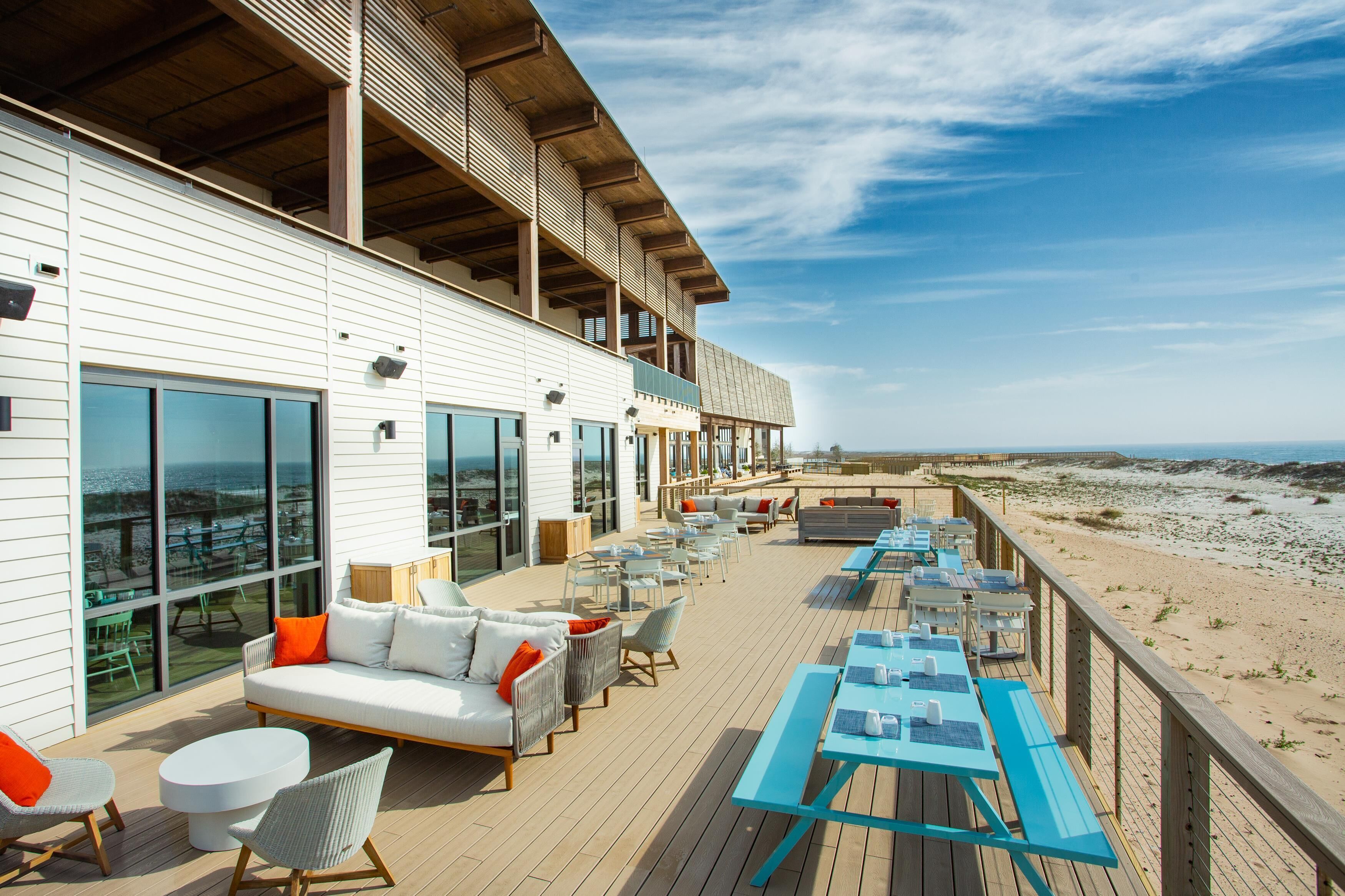 A deck with outdoor seating, a view of the beach, and a building with large windows.