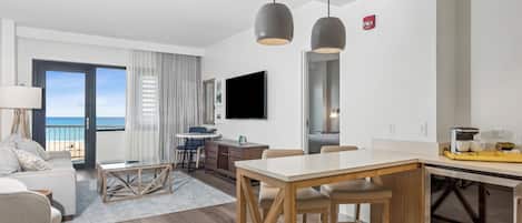 A modern hotel room with a dining area, a TV mounted on the wall, and a view of the beach.