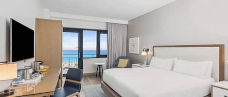 Quarto de hotel com cama, escrivaninha, cadeira, televisão e vista para o oceano.