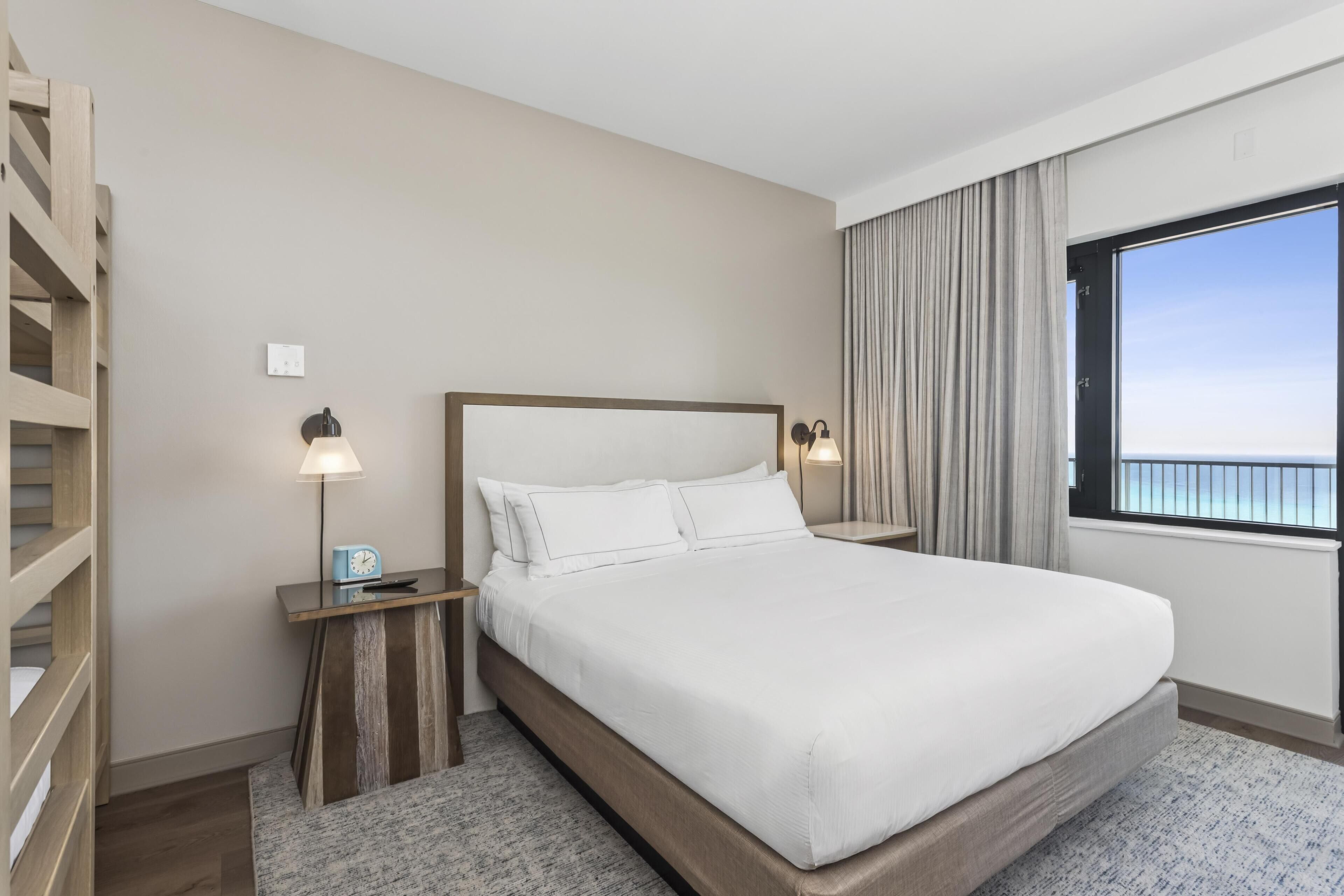 Une chambre d’hôtel dotée d’un grand lit, d’une tête de lit en bois, de tables de chevet avec des lampes, d’une vue sur la mer par une grande fenêtre et d’un balcon.