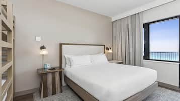 Camera d'albergo con un ampio letto, testiera in legno, comodini con lampade, vista mare attraverso una grande finestra e un balcone.