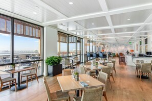 Ein modernes Restaurant mit hohen Decken, großen Fenstern und Blick auf die Stadt. Tische und Stühle sind ordentlich angerichtet, und im Hintergrund befindet sich eine Bar.