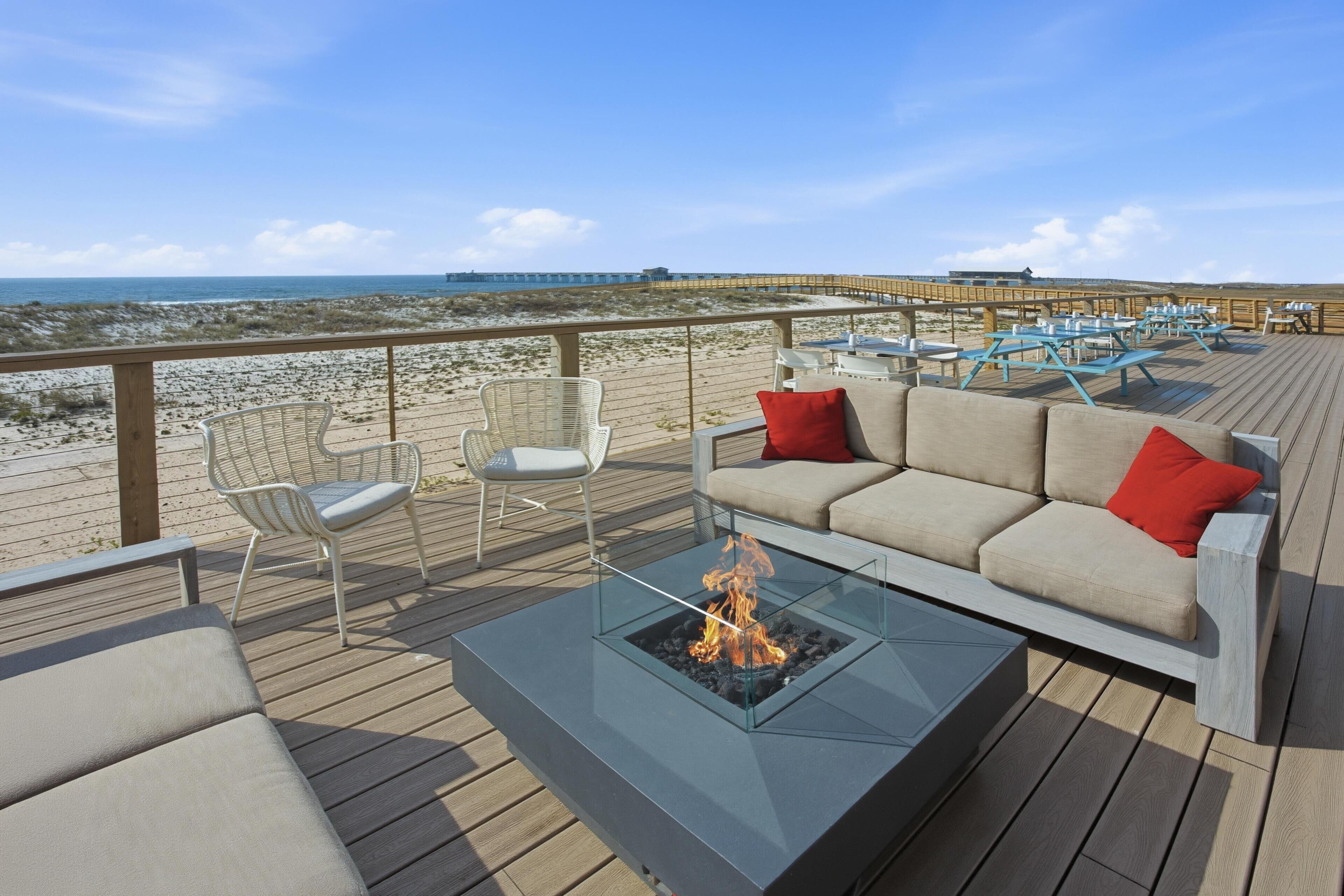 A deck with a glass-top table, a sofa, and chairs, overlooking a beach and pier.