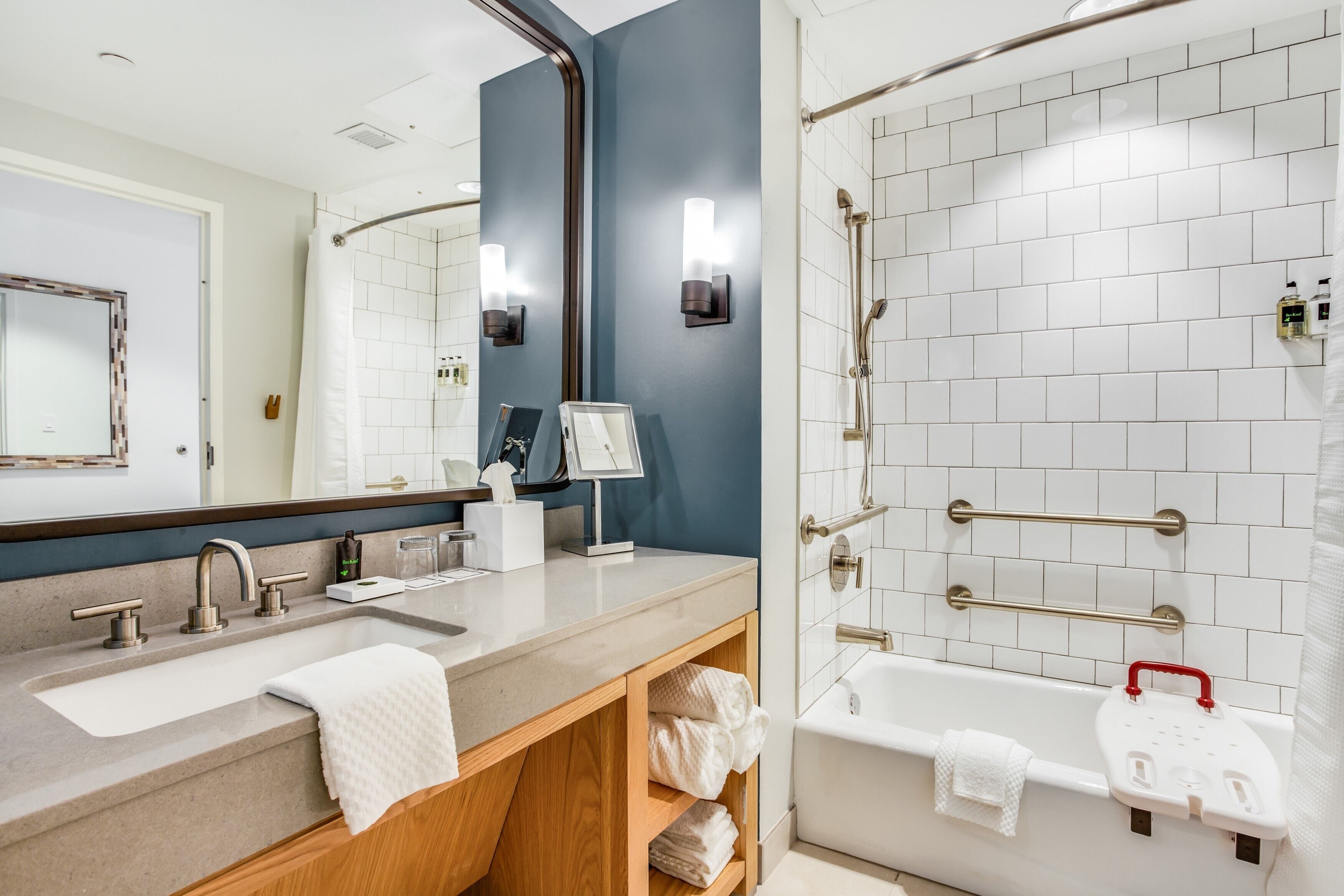 A modern bathroom with a large mirror, dual sinks, a bathtub with grab bars, and a shower with a glass door.