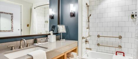 A modern bathroom with a large mirror, dual sinks, a bathtub with grab bars, and a shower with a glass door.