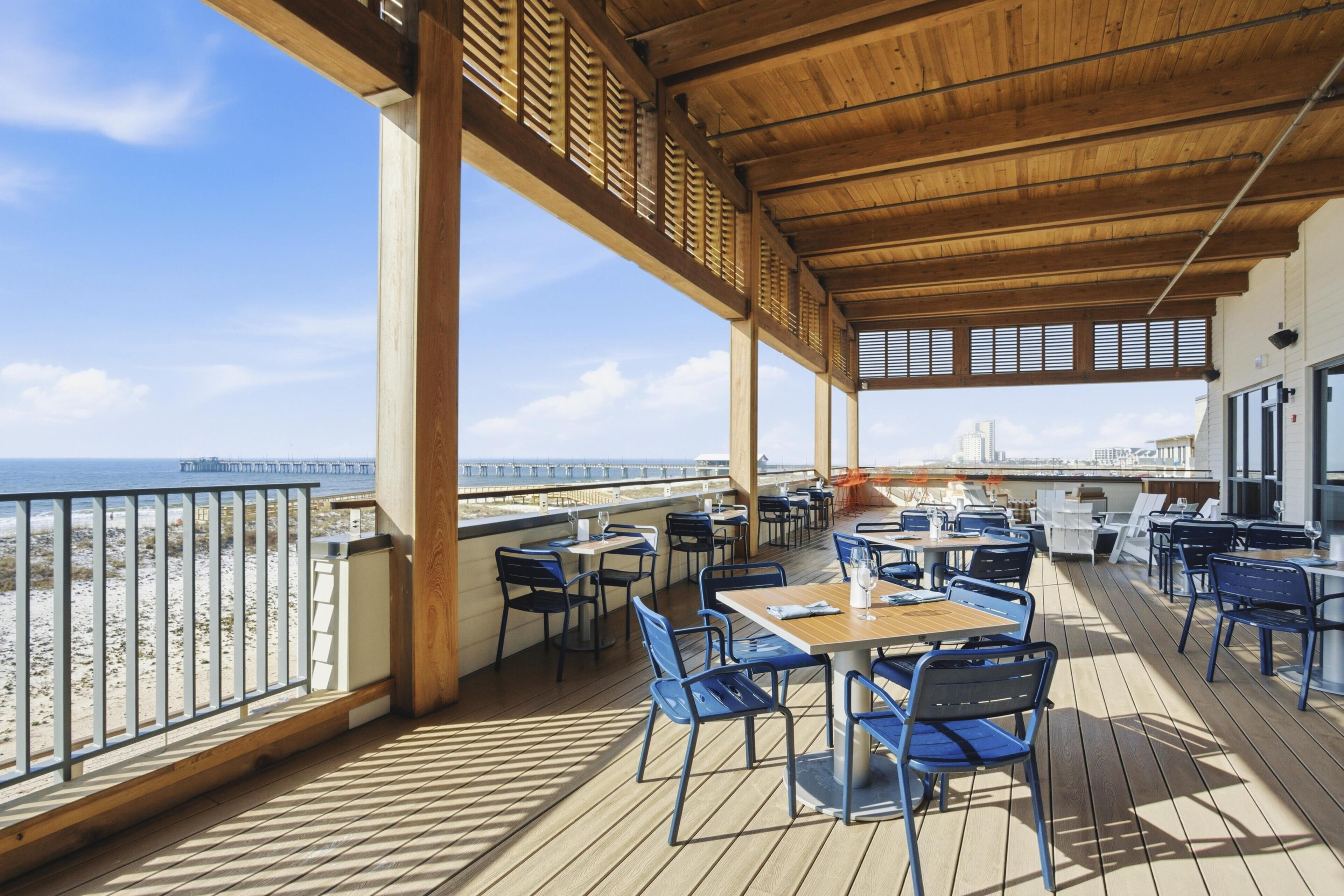 Un espace extérieur abrité, aménagé avec des tables et des chaises, offrant une vue sur une plage et une jetée.