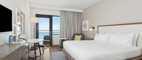 A hotel room with a large bed, a desk, a chair, a small table, a lamp, and a view of the outdoors.