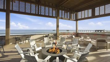 A covered outdoor seating area with a fire pit, white chairs, and a view of the ocean.