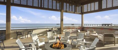 A covered outdoor seating area with a fire pit, white chairs, and a view of the ocean.