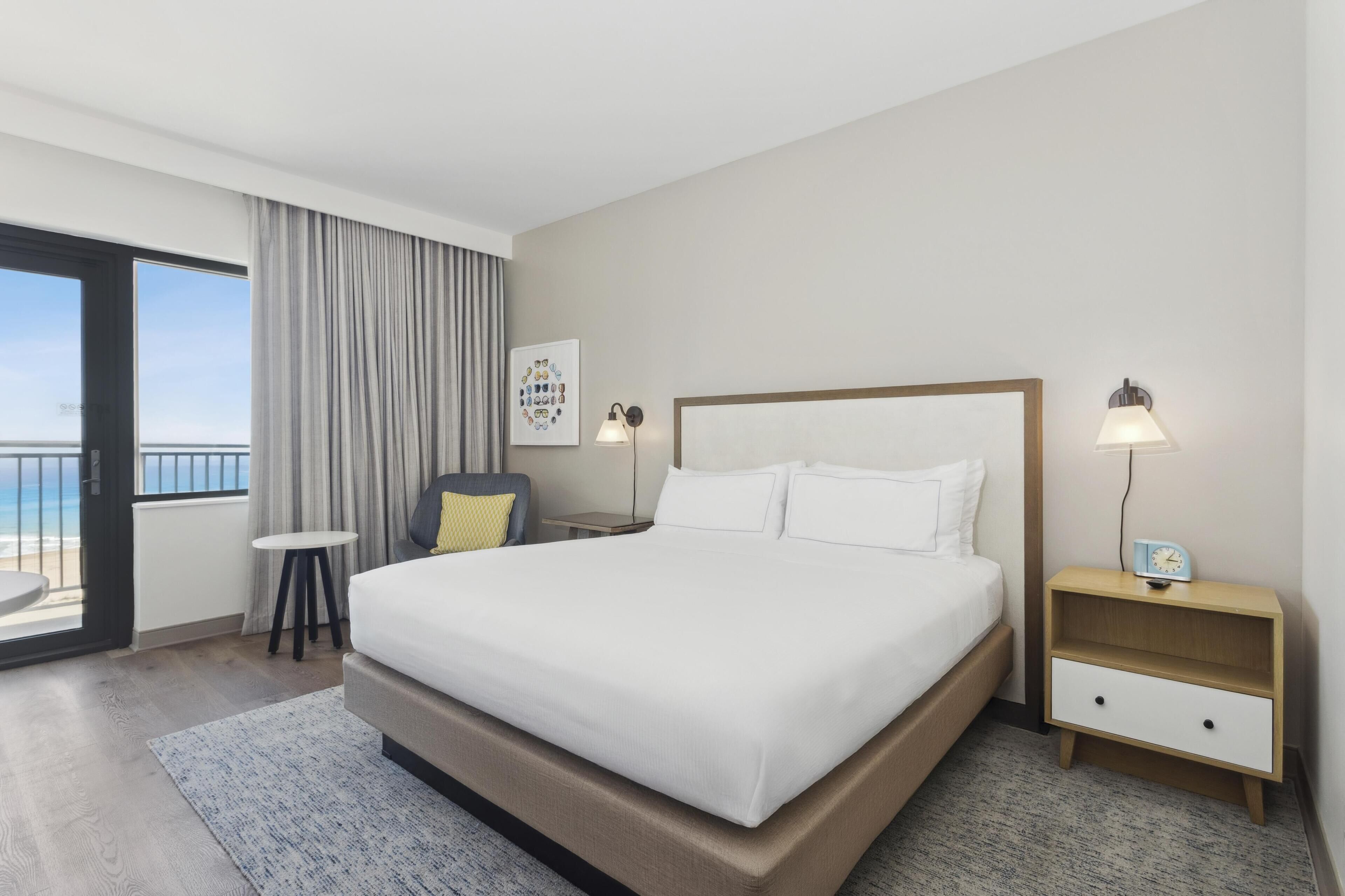 A hotel room with a large bed, a bedside table with a lamp, a small table, and a chair. There is a window with curtains and a view of the ocean.