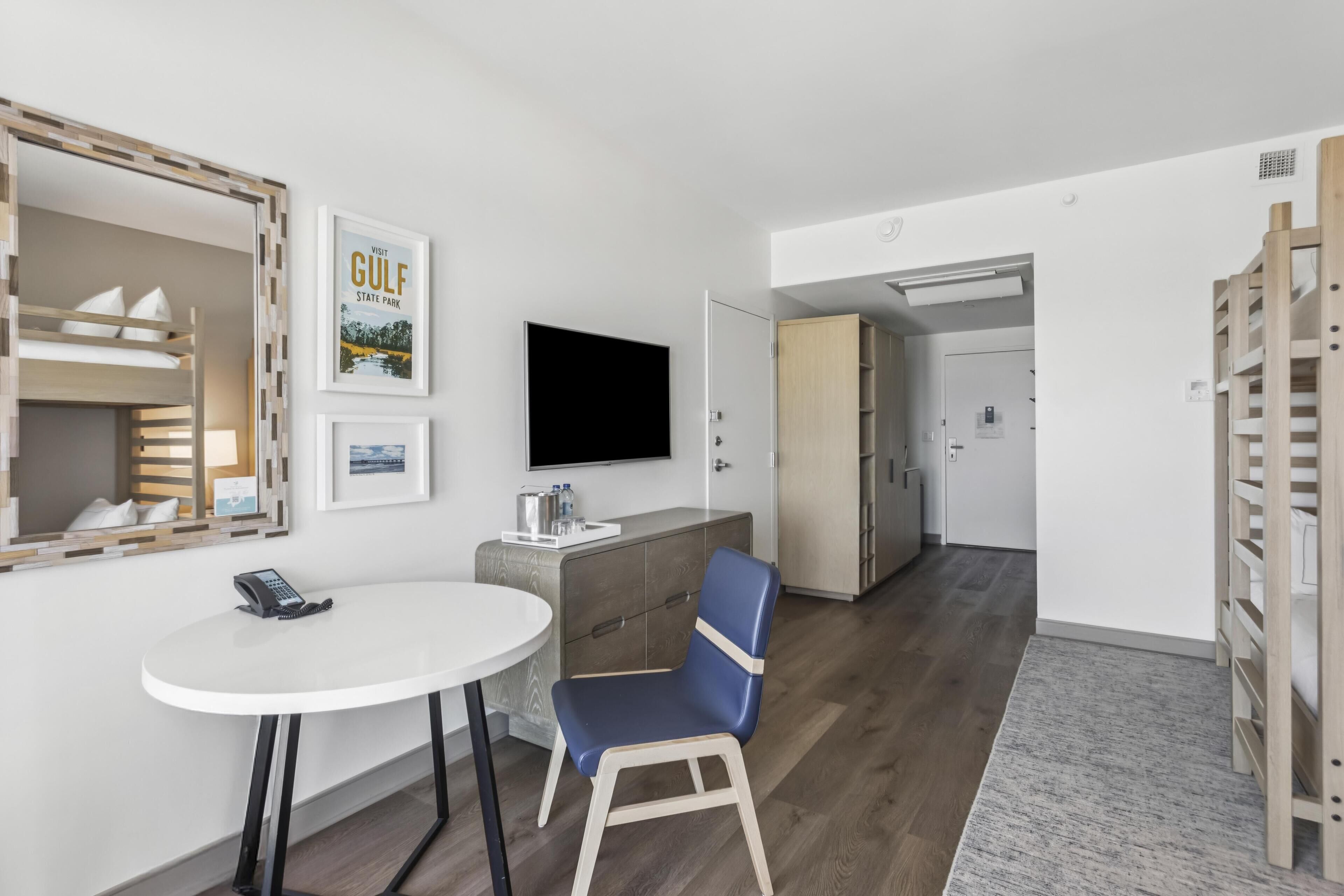 A modern hotel room with a bunk bed, a desk with a phone, a blue chair, a flat-screen TV, and a door leading to a bathroom.