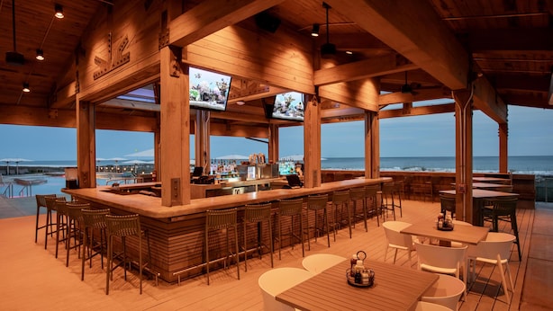 Un bar en plein air avec une terrasse en bois, des sièges, un comptoir et un écran de télévision.