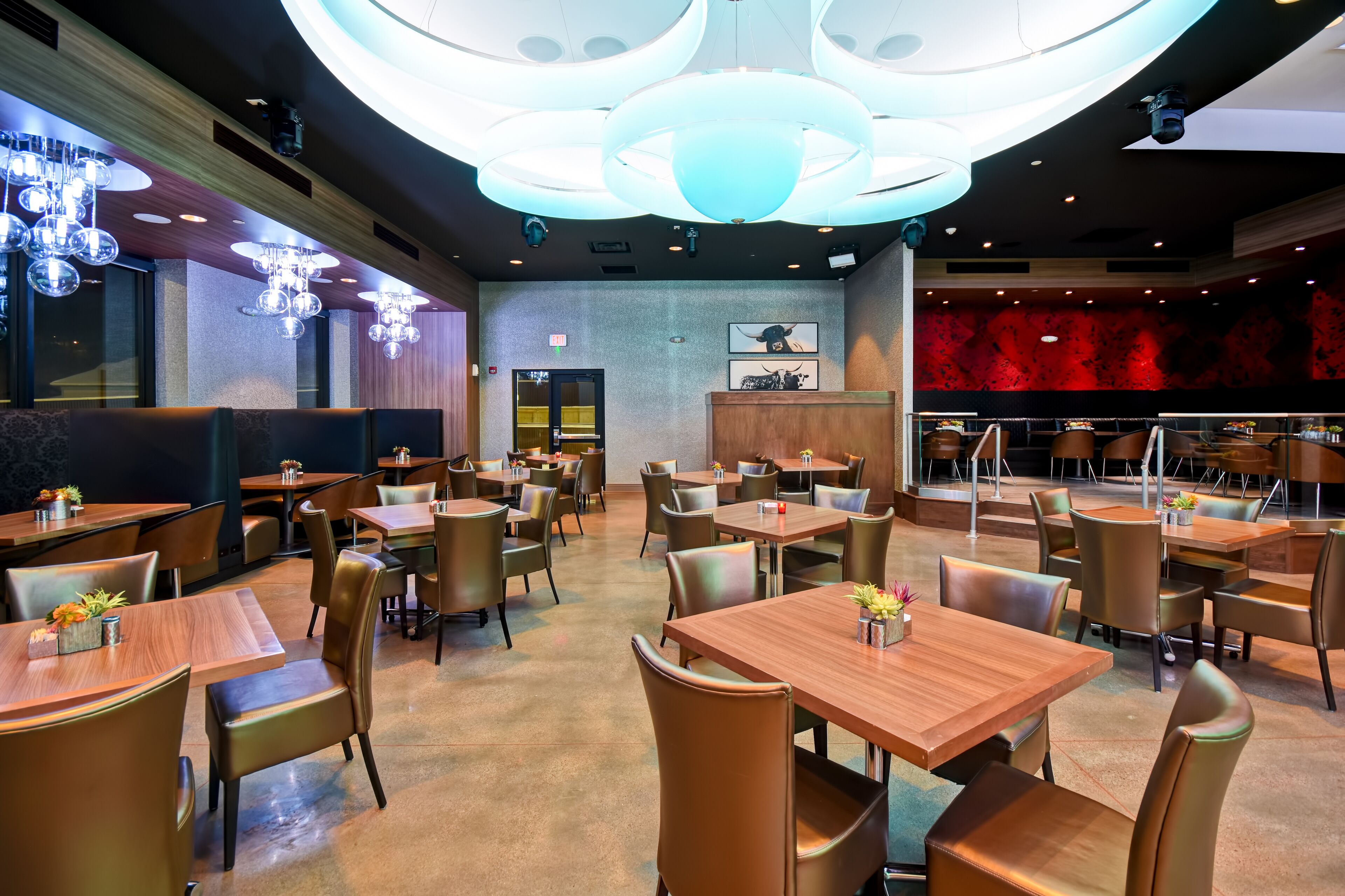 A modern restaurant interior with wooden tables and chairs, a central chandelier, and a red accent wall.