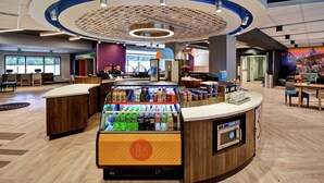 Un café moderne avec un comptoir central, un présentoir réfrigéré, un espace salon et une fresque murale.