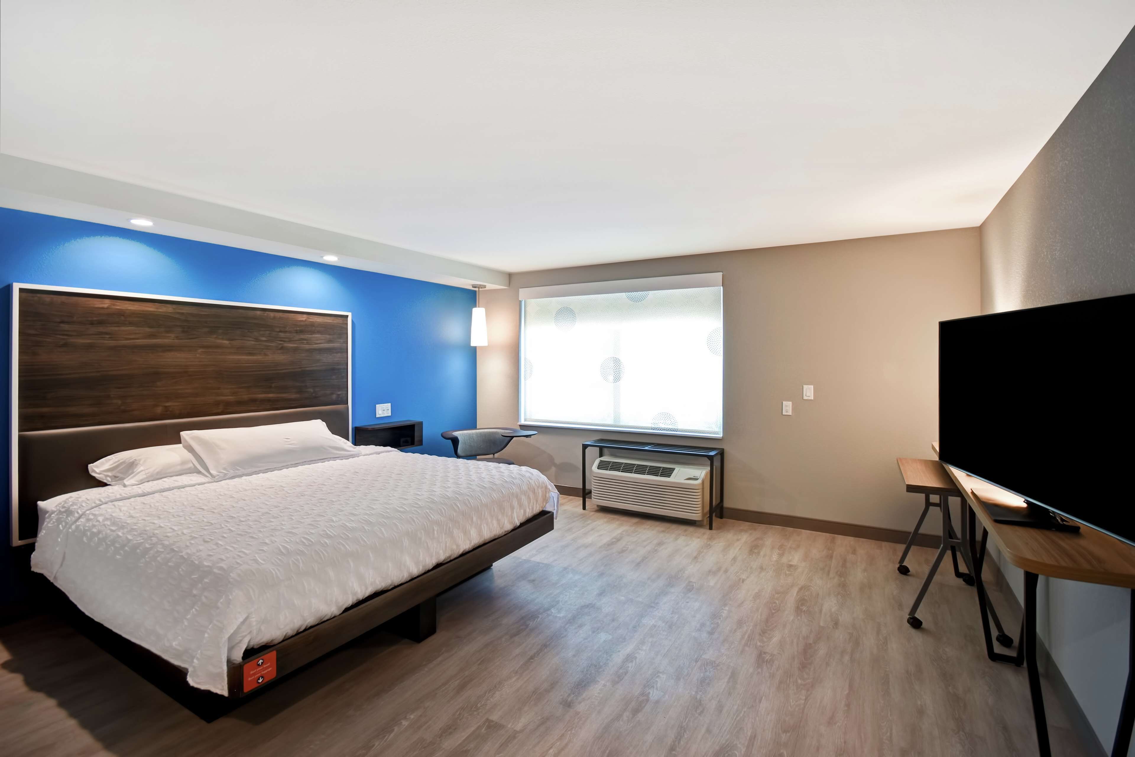 A modern hotel room with a large bed, a flat-screen TV, a desk, and a window with blinds.