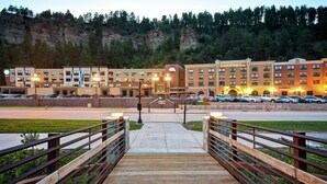 L’entrée d’un hôtel, avec une passerelle en bois menant à un parking, le tout entouré d’un jardin aménagé et avec une chaîne de montagnes en arrière-plan.