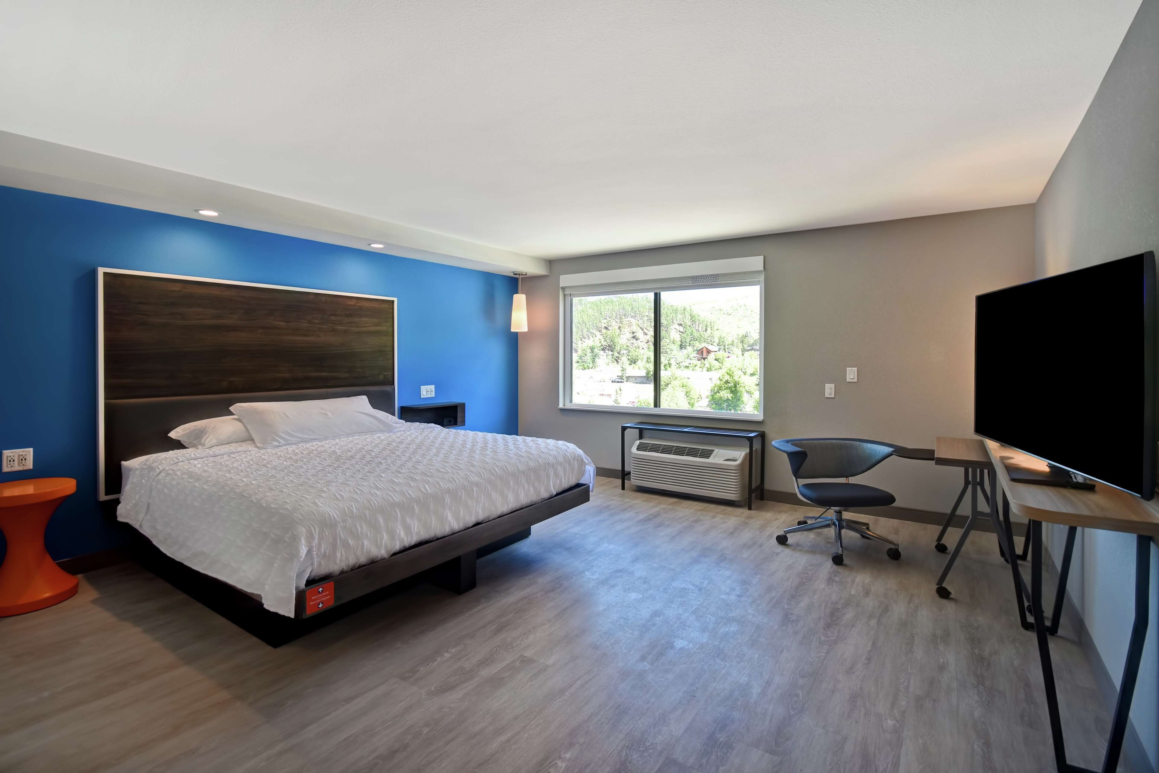 A modern bedroom with a large bed, a desk, a chair, a TV, and a window with a view of greenery.