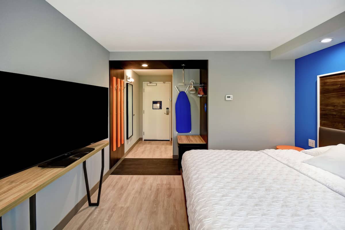 Een hotelkamer met een bed, een televisie op een standaard, een deur en een gang naar een andere kamer.