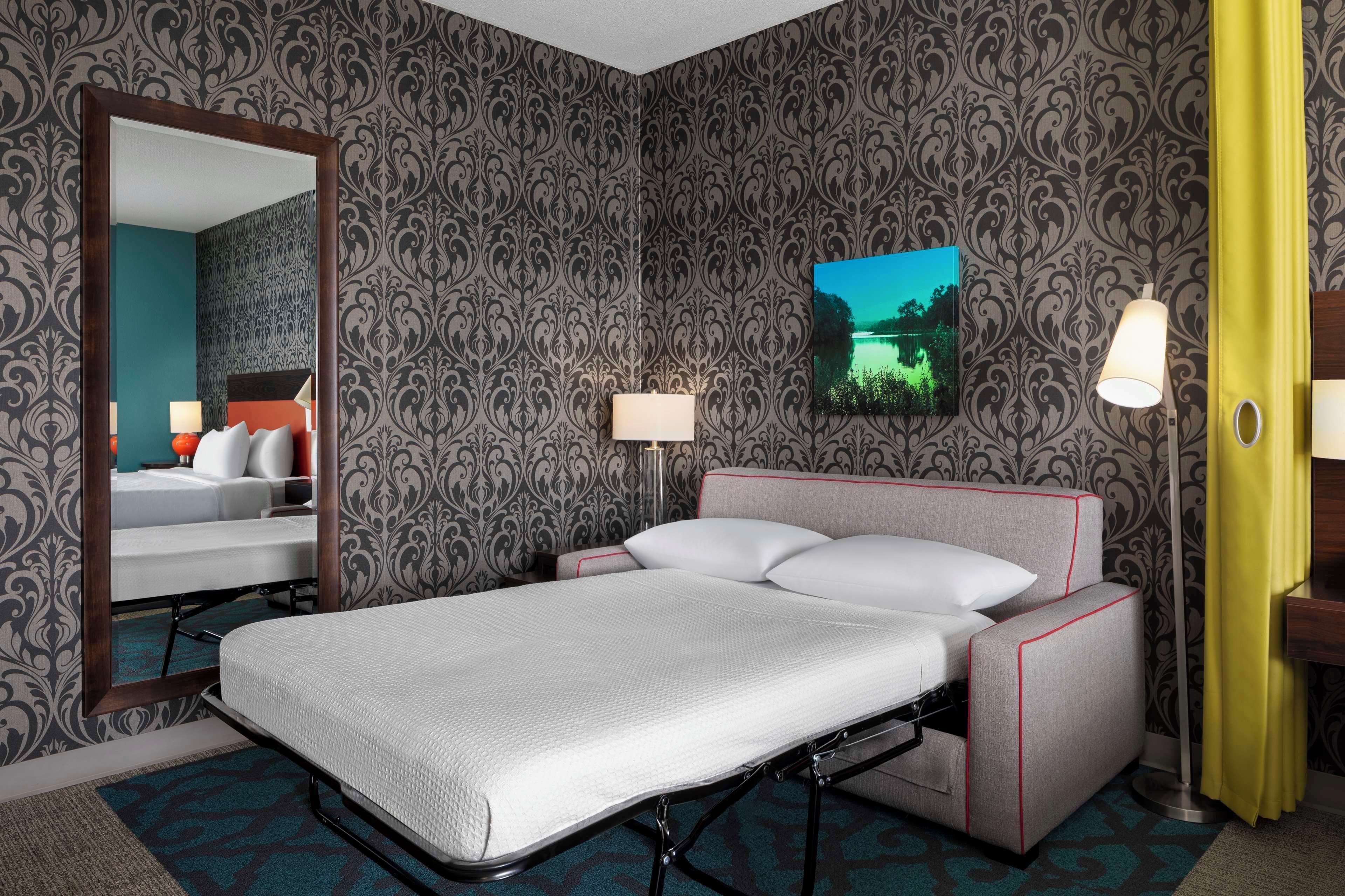 Quarto de hotel com cama, espelho, mesa de cabeceira, abajur e um quadro na parede.