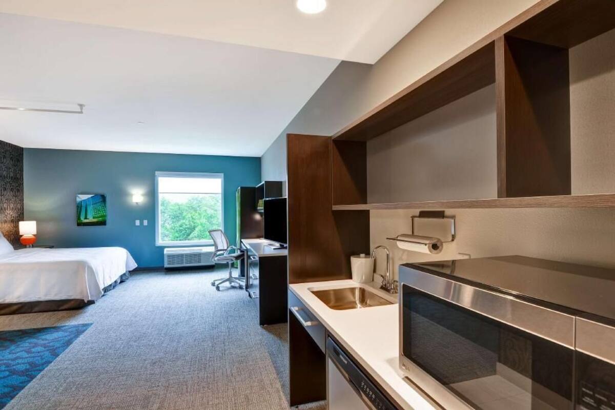 A hotel room with a bed, a desk, a chair, a window, a kitchenette, and a bathroom.
