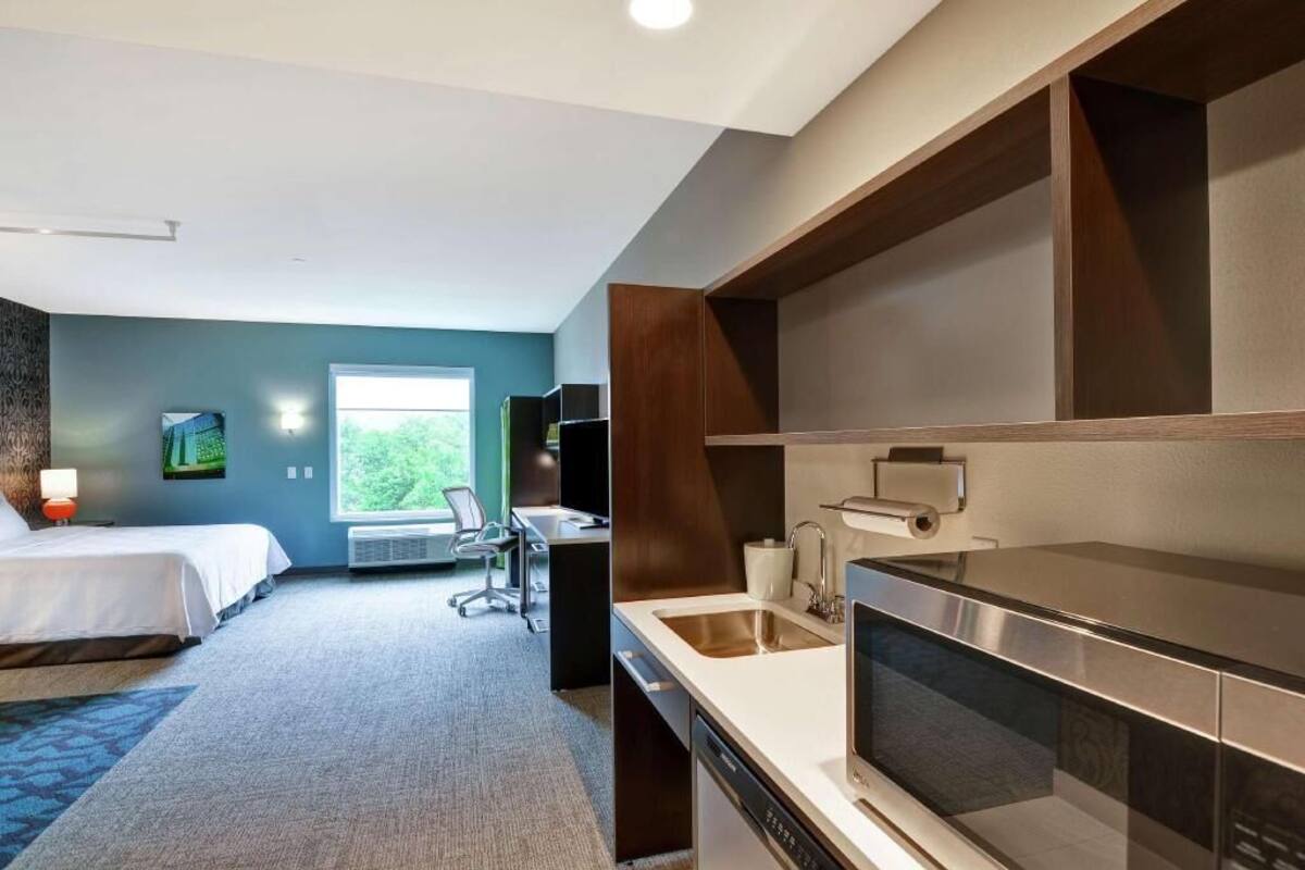 A hotel room with a bed, desk, kitchenette, and a view of greenery.