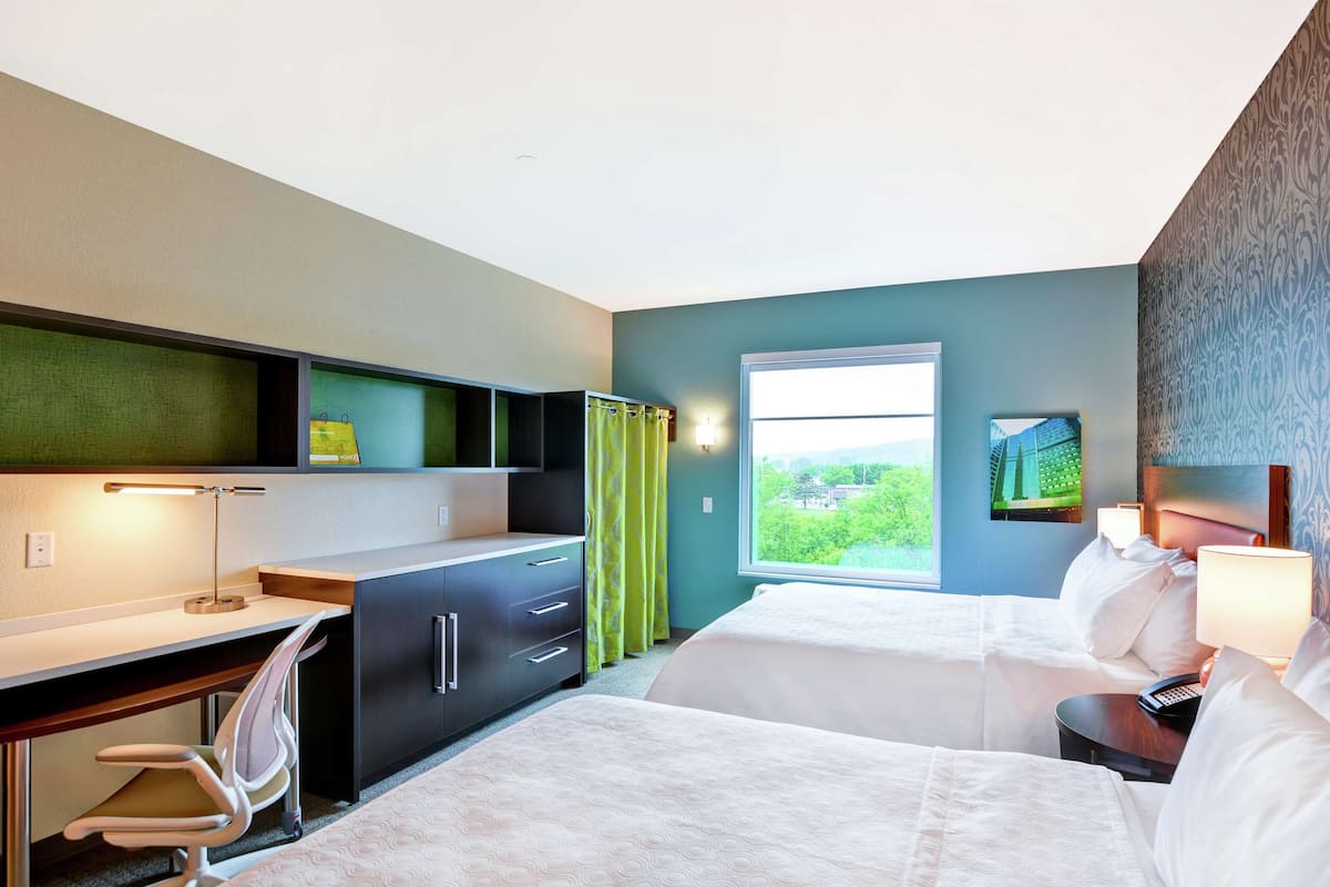 A hotel room with a desk, two beds, a chair, a window with a view, and a painting on the wall.