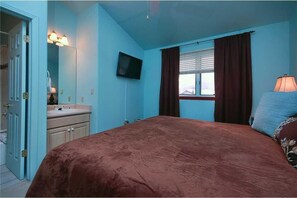 Un dormitorio con una cama grande, televisor, ventana con cortinas y un baño con lavamanos y espejo.