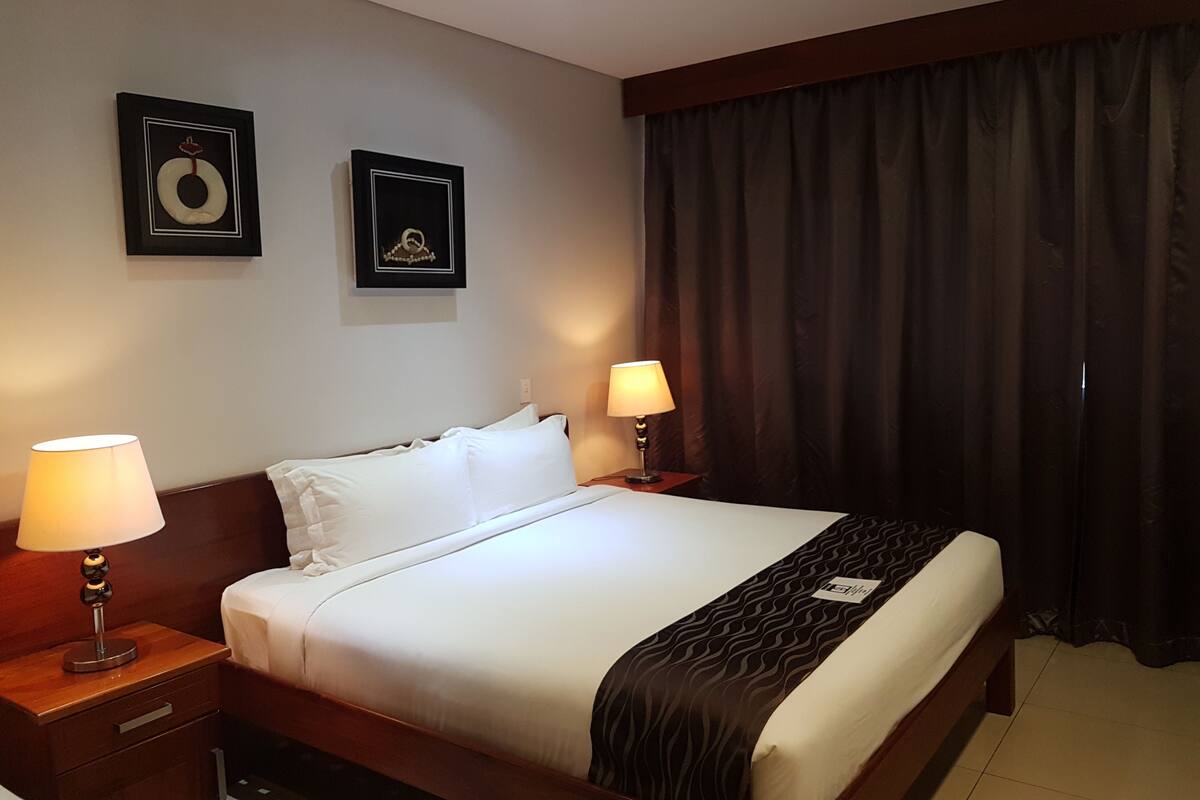 Hotelkamer met een groot bed, twee nachtkastjes, een hoofdeinde, een raam met donkere gordijnen en twee ingelijste foto's aan de muur.