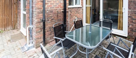 A patio with a glass table and metal chairs, a black umbrella, and a brick wall.