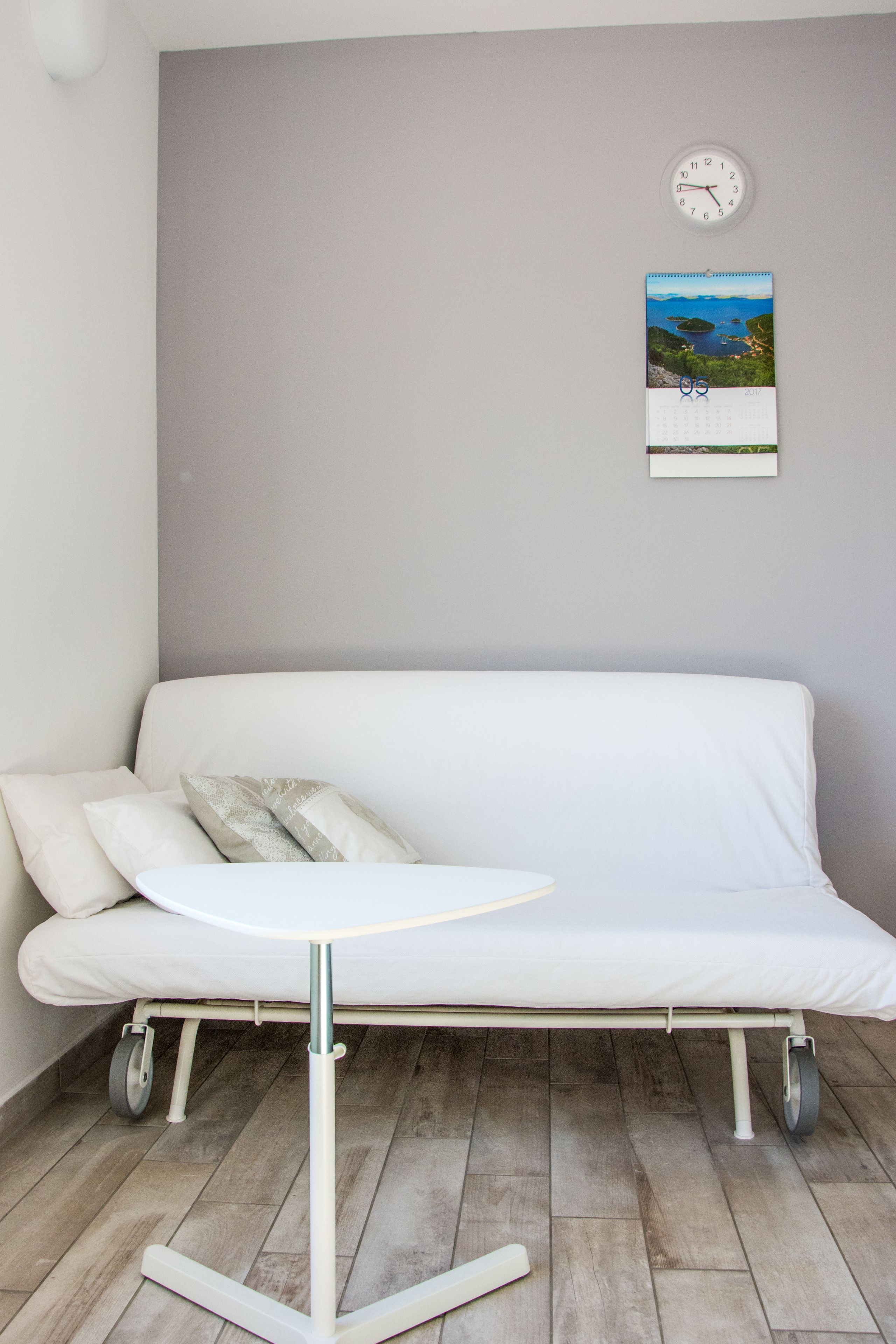A white sofa with a small table and wheels, against a grey wall with a clock and a calendar.