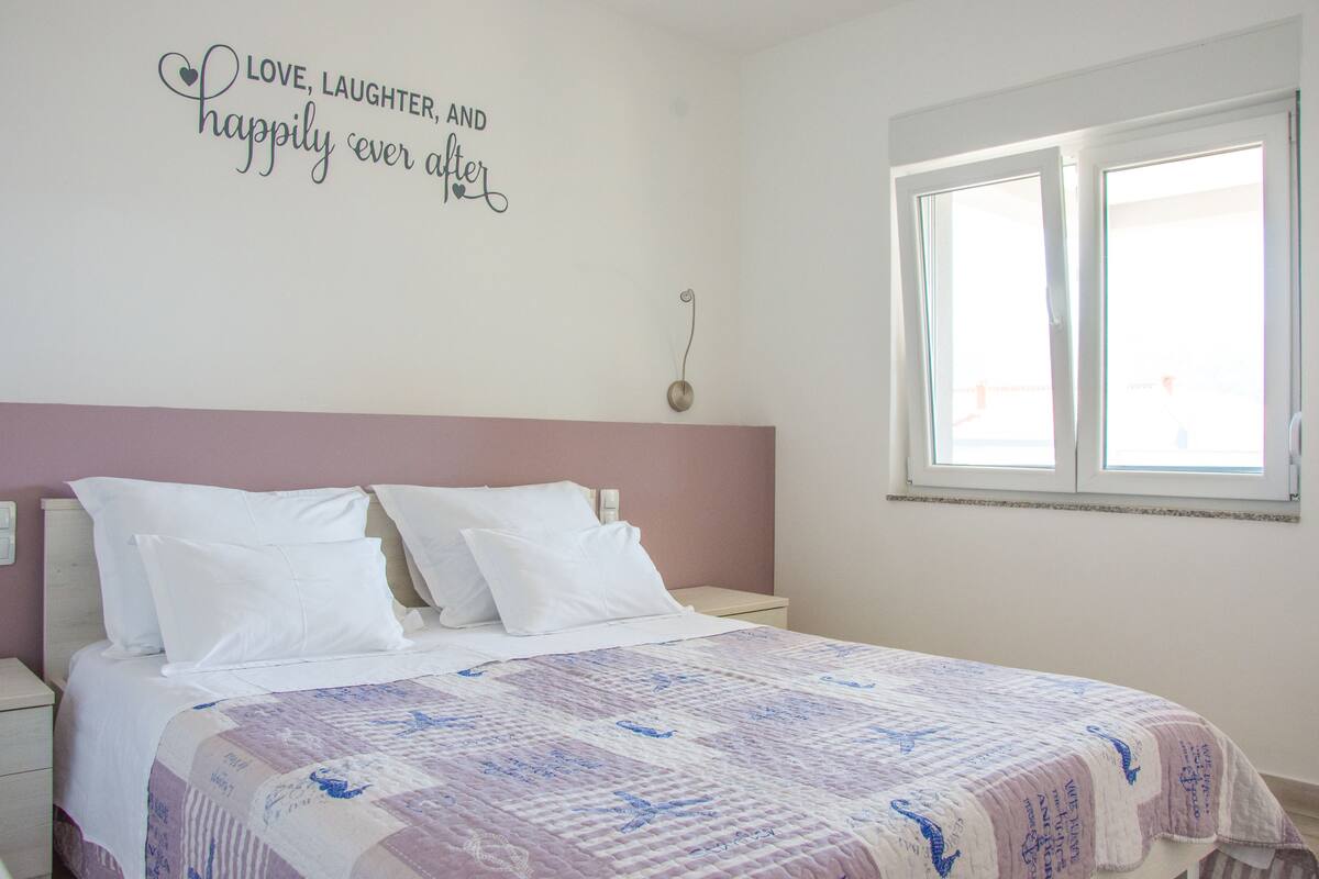 Un dormitorio con una cama, almohadas blancas, colcha, decoración en la pared, mesita de noche y una ventana.