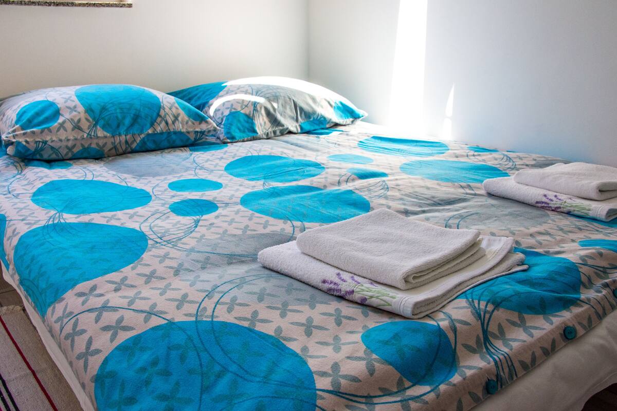 Una cama bien hecha con ropa de cama de diseño azul y blanco, una toalla blanca doblada y una ventana que proporciona luz natural.