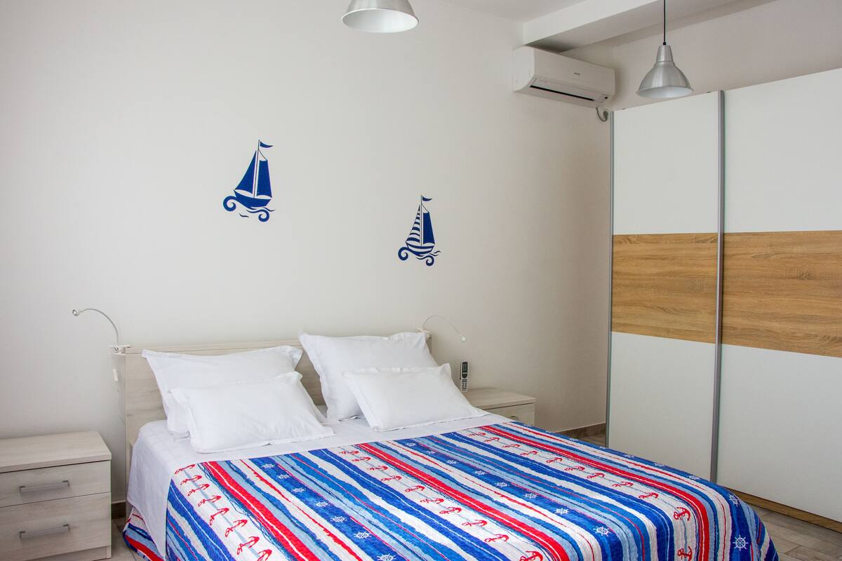 Un dormitorio con cama, mesitas de noche blancas, cabecero de madera y una pared decorada con calcomanías de veleros.