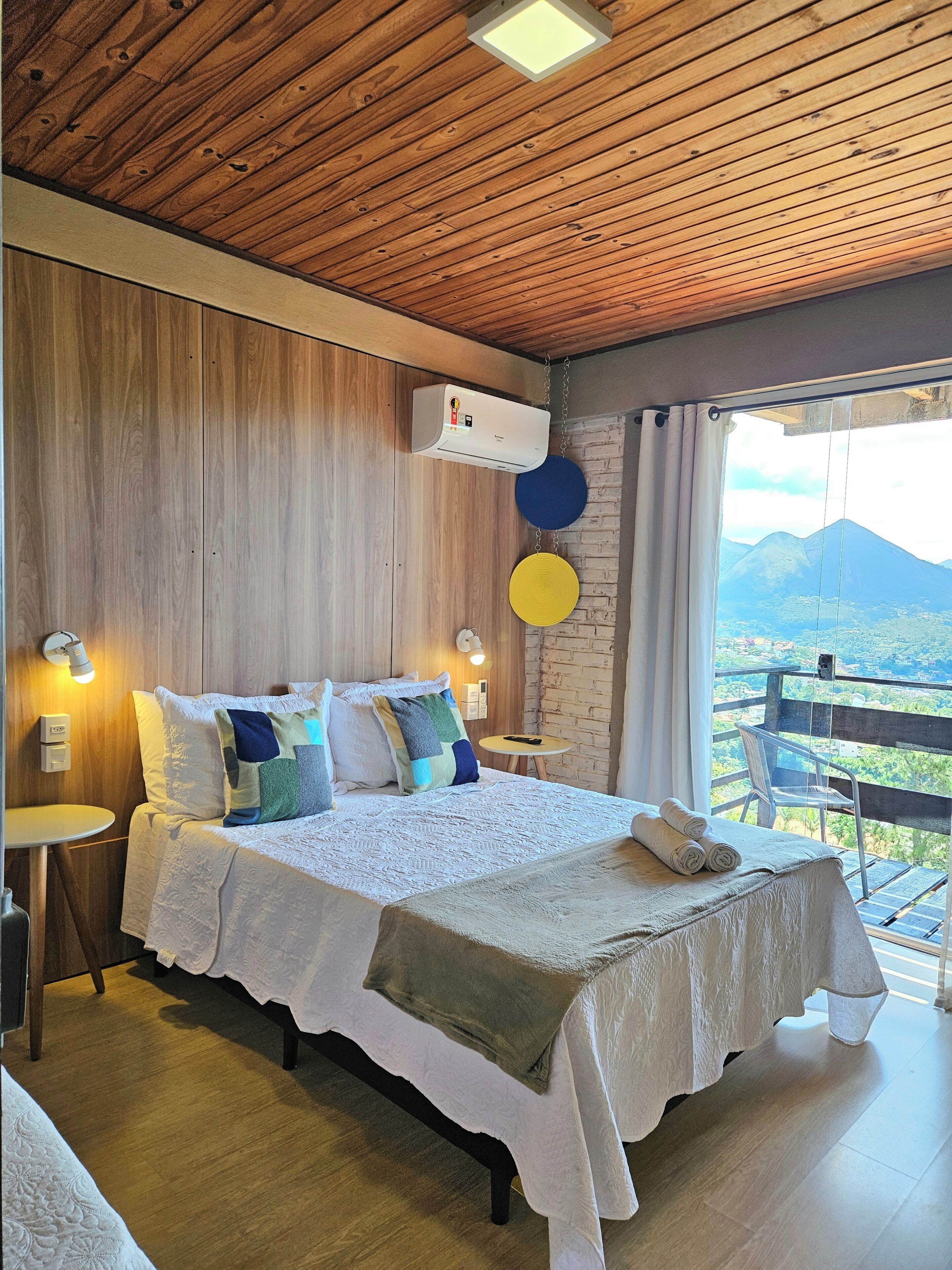 A bedroom with a wooden ceiling, a bed with white bedding, a small table, and a view of mountains.