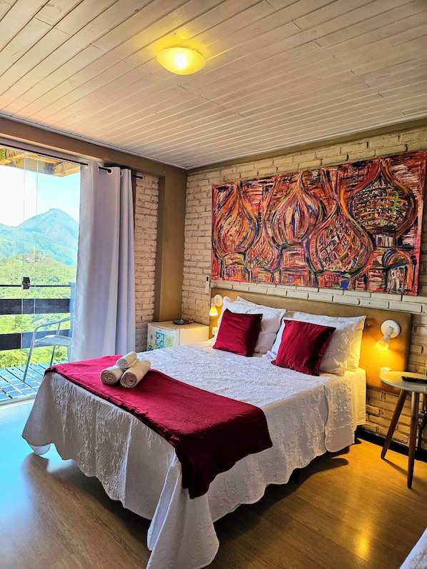 A bedroom with a bed, a large window, a brick wall, and a colorful artwork.