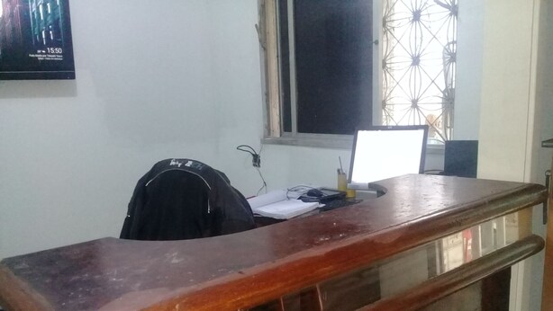 Un escritorio de madera con una mampara de vidrio, una chaqueta negra en el respaldo, un cartel en la pared y una ventana con una celosía decorativa.