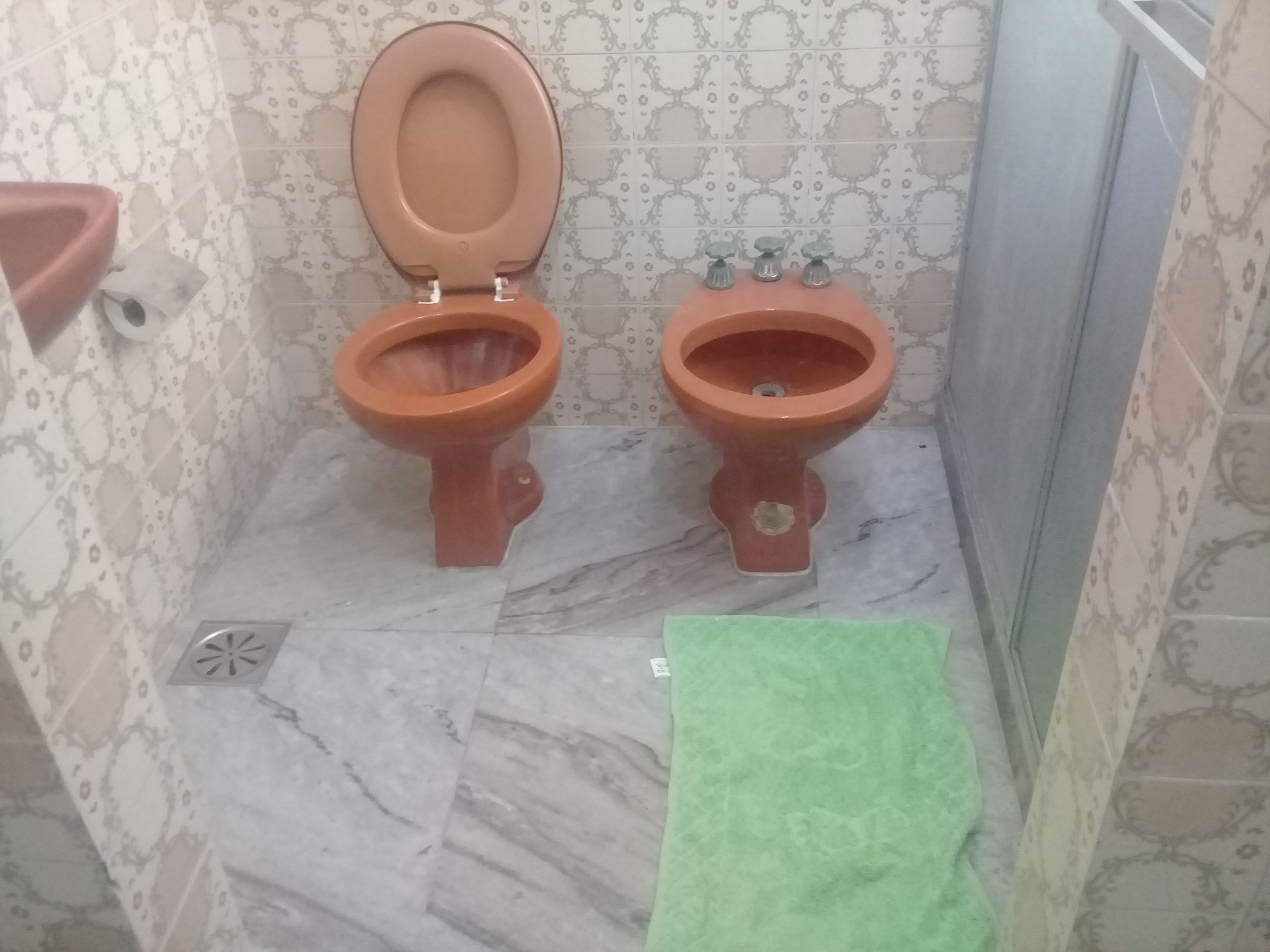 Un baño con dos inodoros, piso de mármol, papel tapiz estampado y una toalla verde.