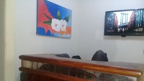 Ein Zimmer mit einem hölzernen Empfangsschalter, einem farbenfrohen Bild an der Wand und einem Fernseher, der eine Stadtansicht zeigt.