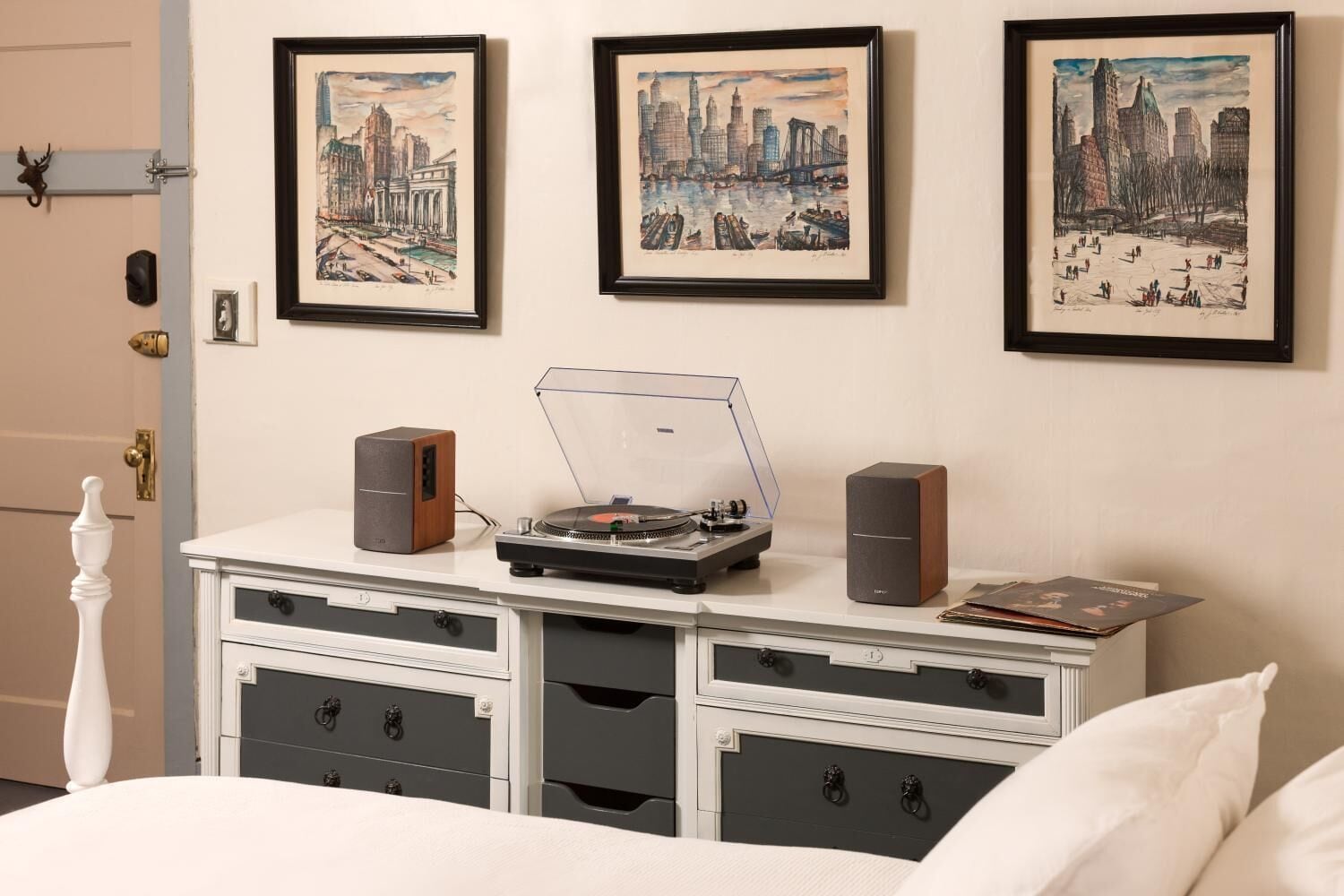 A room with a bed, a dresser with drawers, a record player, speakers, and framed cityscape pictures on the wall.