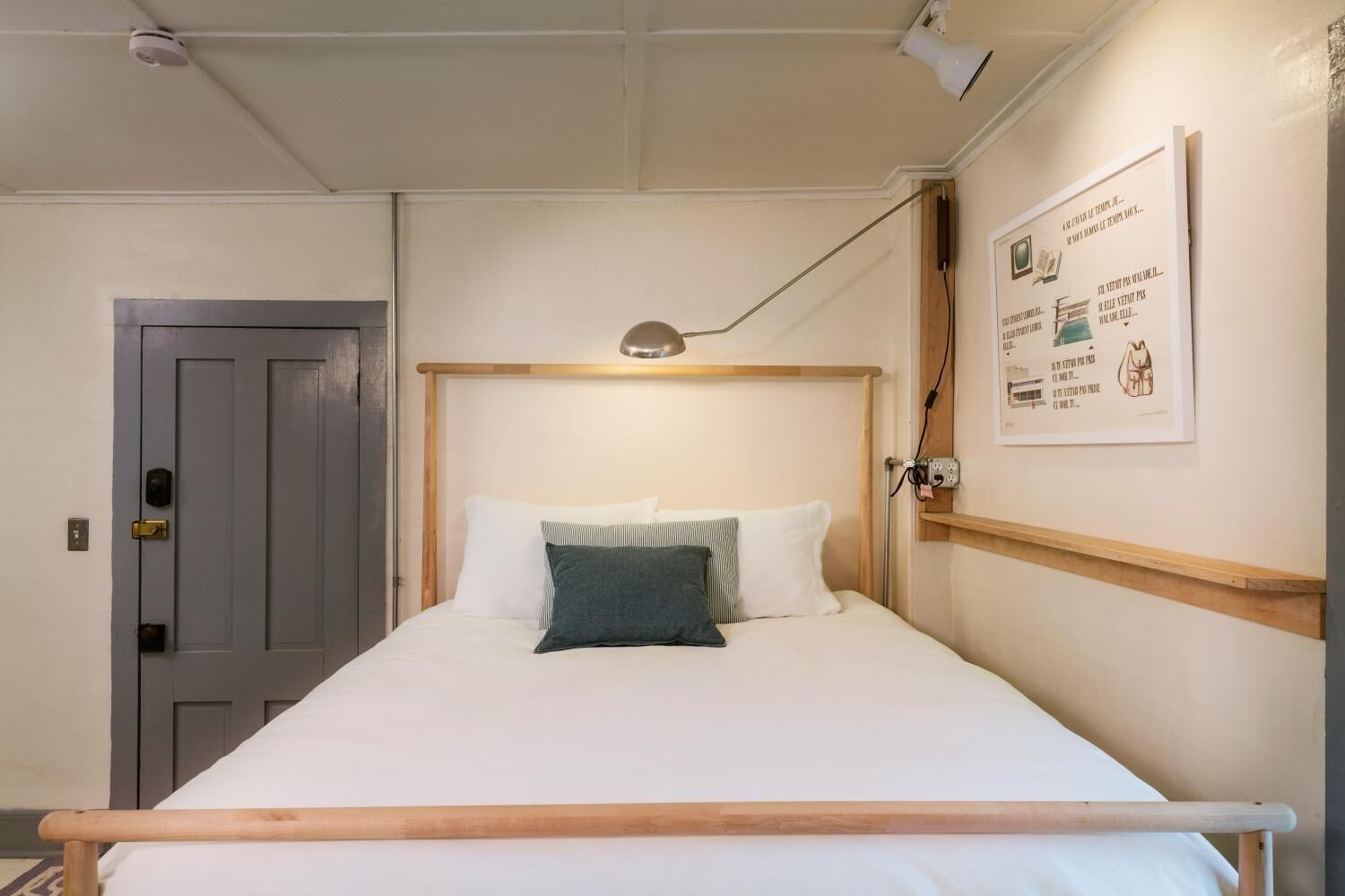Una cama individual con cabecera de madera, ropa de cama blanca y almohada verde. Hay una lámpara de noche, una puerta con cerradura y un aplique de pared.