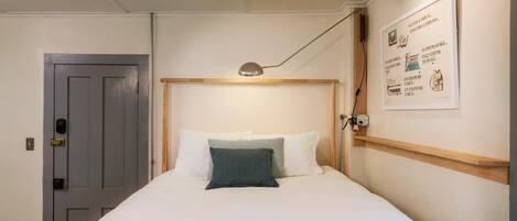 Una cama individual con cabecera de madera, ropa de cama blanca y almohada verde. Hay una lámpara de noche, una puerta con cerradura y un aplique de pared.