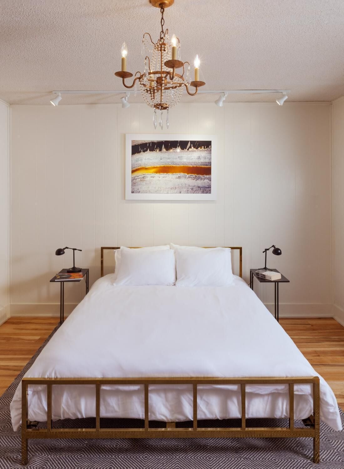 Une chambre à coucher avec un lit en bois, du linge de lit blanc, un lustre, un tableau encadré au mur et deux tables de chevet avec des lampes.
