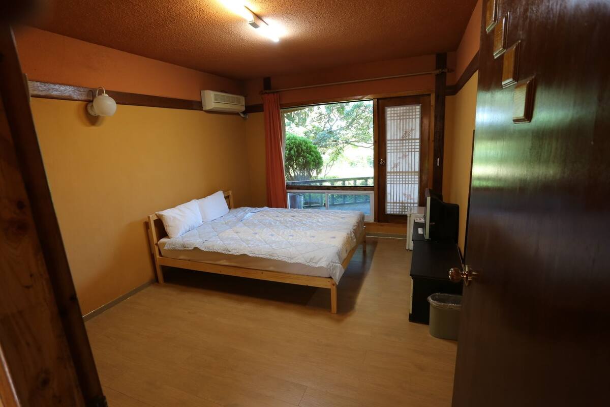 A room with a bed, a wooden floor, a ceiling fan, and a window with a view of trees.