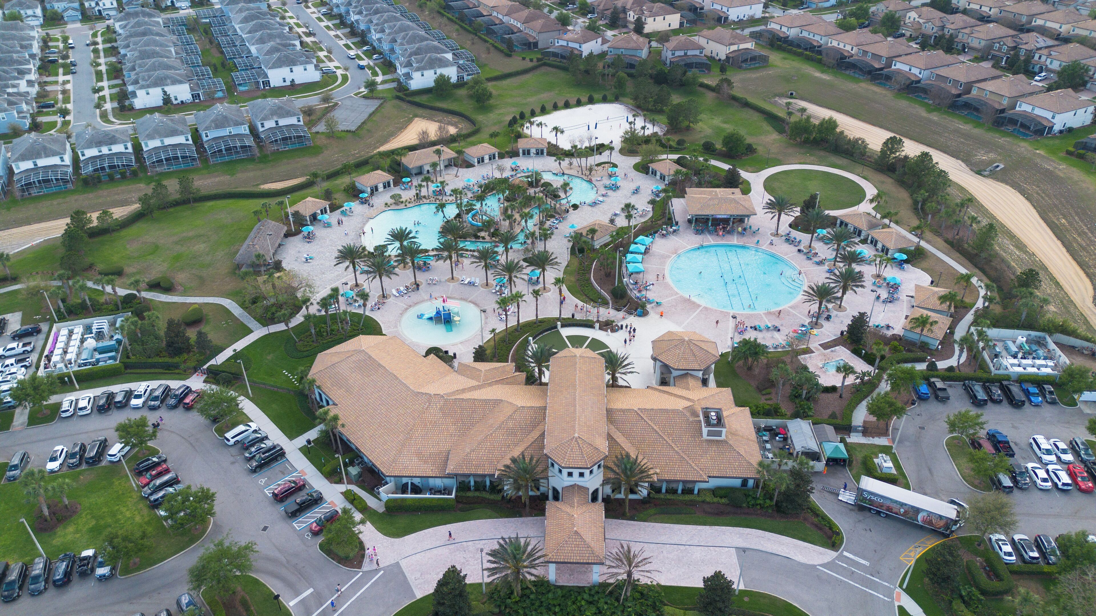 Vista aérea de un resort con varias piscinas, un edificio central con techo de tejas y un estacionamiento con varios automóviles.
