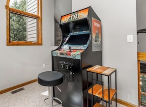 A retro arcade machine labeled "Tetris" in a room with a window, a stool, and a small wooden table. - Unwind at High Ridge: Hot Tub Nights, Arcade Lights & Fire Pit Fun! (Galena)