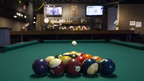 Una mesa de billar con bolas dispuestas en forma de triángulo, una zona de bar con botellas y un televisor al fondo.