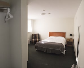 Una habitación de hotel con cama, mesita de noche, silla, lámpara y armario.