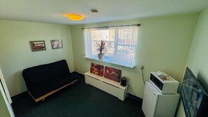 A small, sparsely furnished room with a single bed, a small table, a window with curtains, a microwave, and a television.